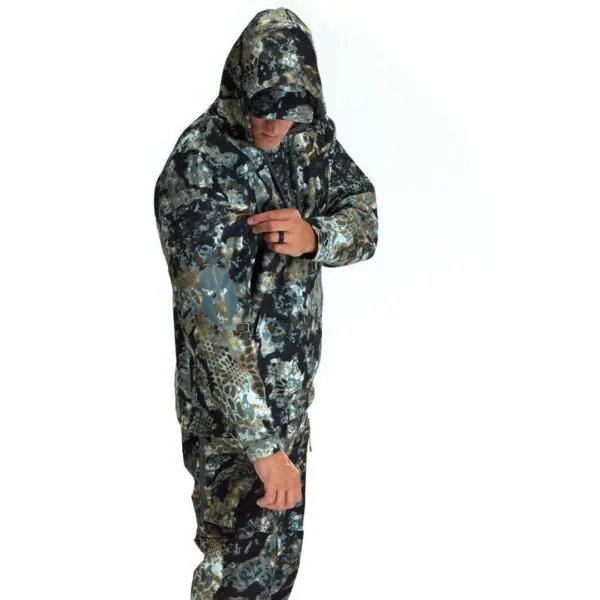 A person wearing a Kryptek Tartaros Hoodie Obskura Transitional, including a hood and cap, faces slightly to the side and adjusts something on their upper left sleeve. The camo pattern includes shades of green, brown, grey, and black. The background is plain white.