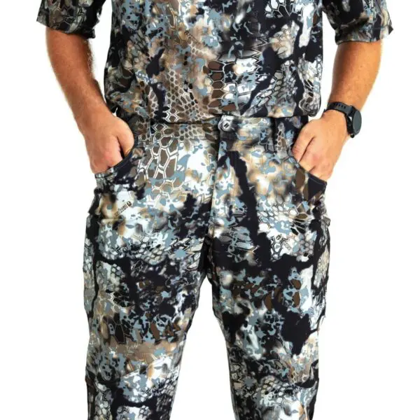A person dressed in Kryptek Valhalla Pants Obskura Transitional stands with their hands in their pockets. The clothing features a mix of black, gray, brown, and white colors. The person also wears a black wristwatch. The background is white.