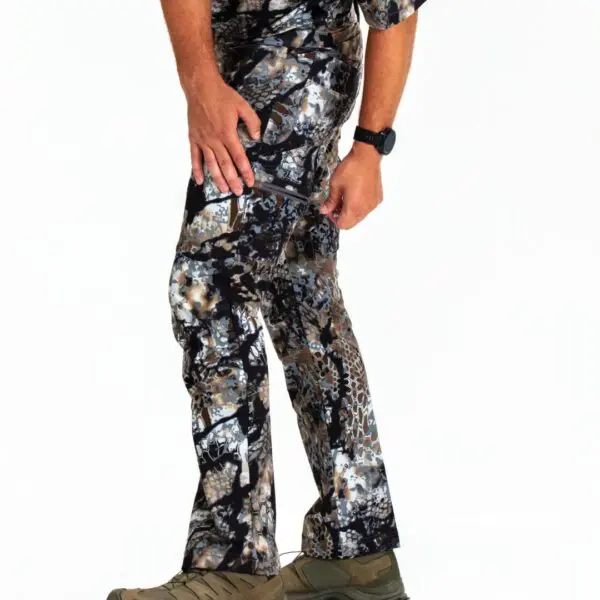 A person wearing Kryptek Valhalla Pants Obskura Transitional, along with a camouflage-patterned top, is shown from the chest down. The person is also wearing a black watch on their left wrist and green outdoor boots. The background is plain white.