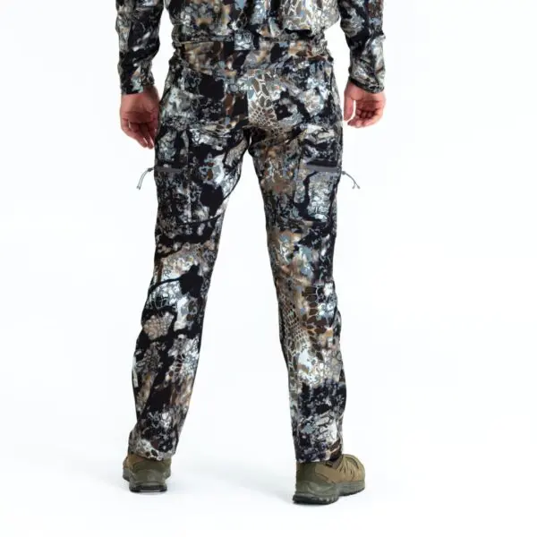 A person stands facing away, wearing a camouflaged outfit consisting of a long-sleeve shirt and matching Kryptek Valhalla Pants Obskura Transitional. The camouflage pattern includes shades of brown, green, and gray. They are also wearing brown hiking shoes. The background is plain white.