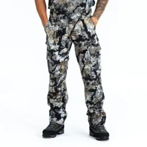 Person wearing Kryptek Stalker Pants Obskura Skyfall and black hiking boots. Their hands are in the pockets of the pants, and they have a tattoo on their visible left arm. The background is plain white.