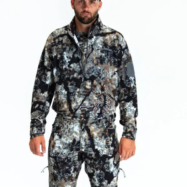 A bearded man stands against a white background wearing a matching Kryptek Valhalla Long Sleeve Zip Highlander outfit consisting of a jacket and pants. The camouflage pattern features shades of blue, brown, and beige. He has a serious expression and his hands are by his sides.