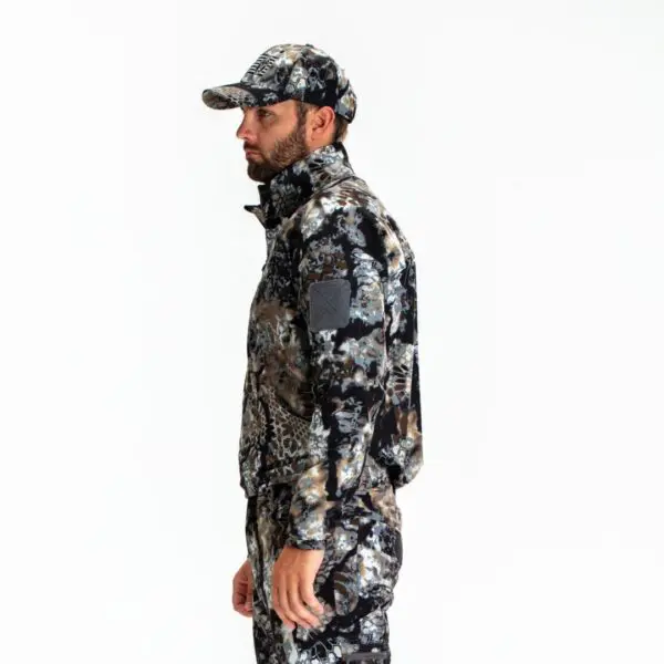 A man stands in profile against a plain white background, wearing a matching hat, jacket, and pants in a camouflaged pattern. His Kryptek Valhalla Long Sleeve Zip Highlander attire appears to be designed for outdoor activities, emphasizing blending in with natural surroundings.