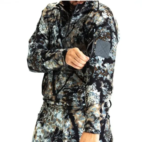 A person is wearing a camouflage-patterned jacket and pants. They are pulling a zipper pocket on the sleeve of the Kryptek Valhalla Long Sleeve Zip Highlander. Their face is not visible in the image.