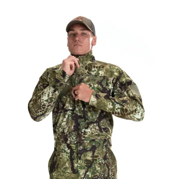A person wearing a cap and a full-body camouflage outfit zips up the collar of their Kryptek Valhalla Long Sleeve Zip Highlander. The background is white.