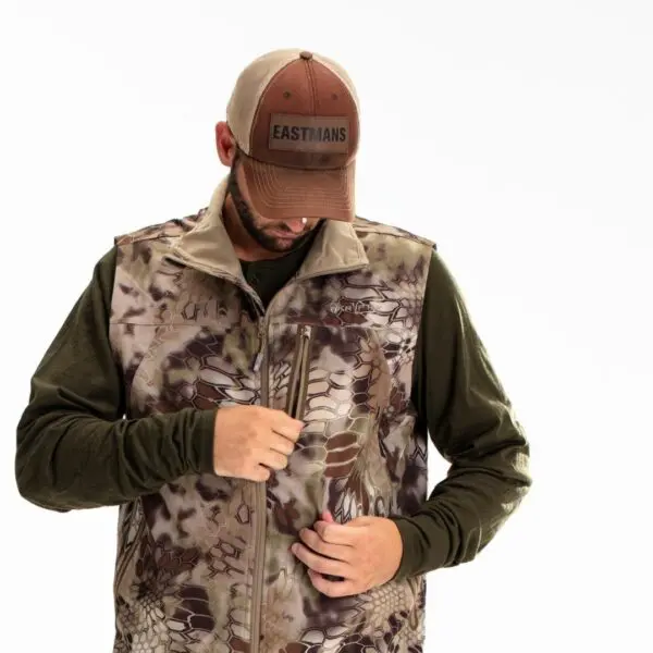A man wearing a Kryptek Dalibor Vest over a green long-sleeve shirt is zipping up his vest. He is also wearing a brown cap with the word "EASTMANS" on it, and his head is tilted downward, focusing on the zipper. The background is plain white.