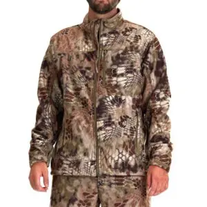 A person stands wearing a Kryptek Njord Windproof Jacket and matching pants. The camo features a mix of brown, green, and beige hues, with reptile-scale-like patterns. The jacket has long sleeves, a front zipper, and a high collar. The face is partially visible.