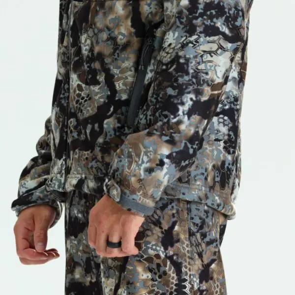 A person is shown from the shoulders down wearing a matching camouflage jacket and pants. The pattern is a combination of various shades of brown, black, white, and grey. The person's hands are visible, and one hand wears a black ring.
