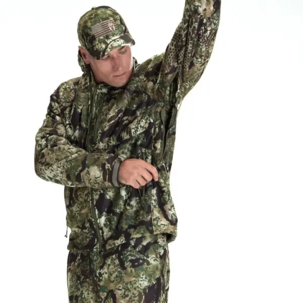 A person wearing a green camouflage outfit, including a Kryptek Njord Windproof Jacket and cap, is shown in a white background. The jacket has a zippered pocket, which the person is accessing with one hand while raising the other arm. The cap features an emblem resembling a flag.
