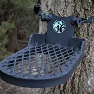 A black Rut Roost is mounted on a tree trunk, secured with a strap. The seat has a grated design, and a logo featuring a silhouetted figure climbing a tree is visible above the seat. Sparse foliage is in the background.