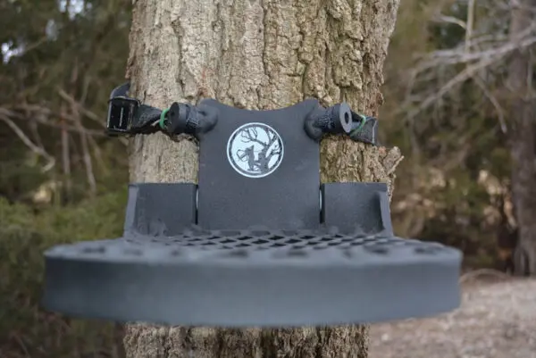 A black Rut Roost hunting tree platform attached to a tree trunk with straps. The platform features a textured surface for grip, and a tree logo is prominently displayed in the center. The background shows a wooded area with dense greenery.