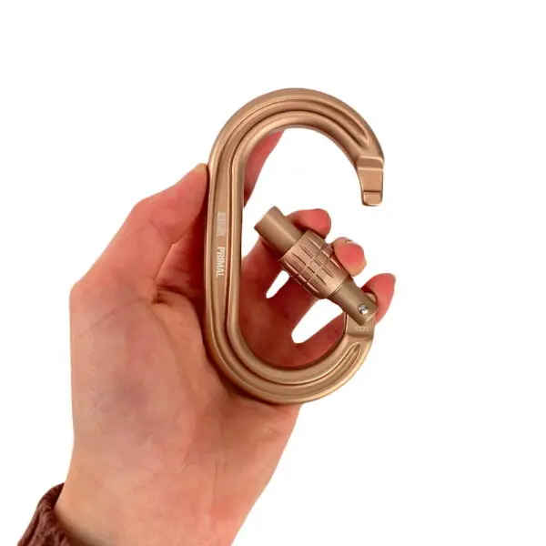 A hand is holding an Ape Canyon Outfitters 8mm Oplux Tether System with Screw Lock Carabiner. The carabiner has a smooth, curved design, and text engraved on its side. The background is plain white, emphasizing the carabiner.