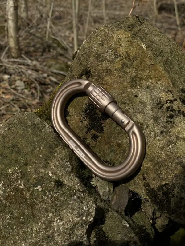An Ape Canyon Outfitters 8mm Oplux Tether System with Screw Lock Carabiner rests against a mossy rock in a forest setting. The background features blurred branches and foliage, suggesting an outdoor adventure or hiking environment.