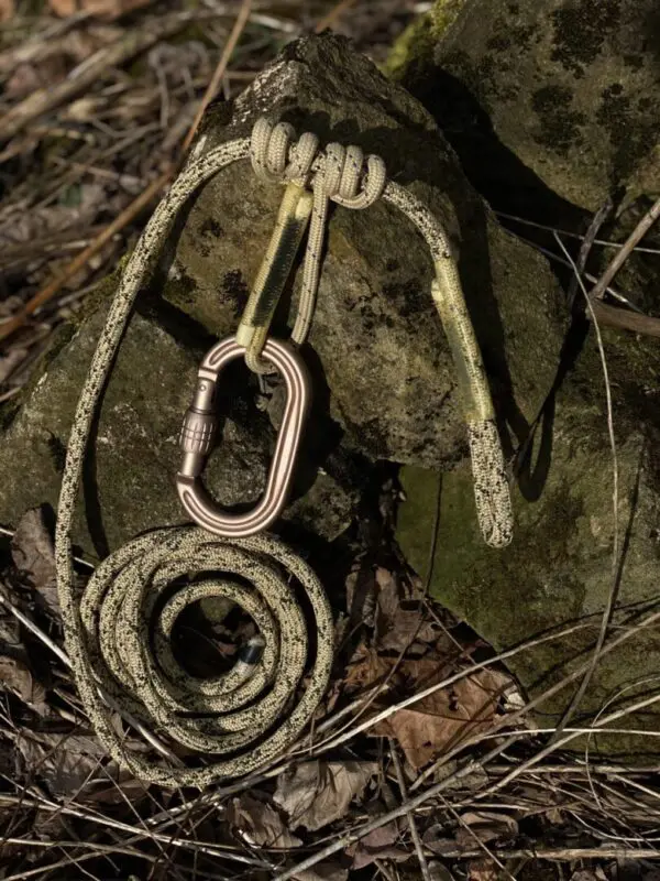 An Ape Canyon Outfitters 8mm Resc Tech Saddle Hunting Combo with Screw Lock Carabiners is placed against a mossy rock and scattered leaves on the ground. The rope and carabiner are light-colored, contrasting with the natural outdoor background.