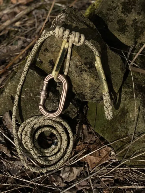 A coiled Ape Canyon Outfitters 8mm Oplux Tether System with Screw Lock Carabiner is placed on moss-covered rocks. The rope and carabiner are both in earthy tones, blending with the natural outdoor surroundings of leaves, twigs, and moss.