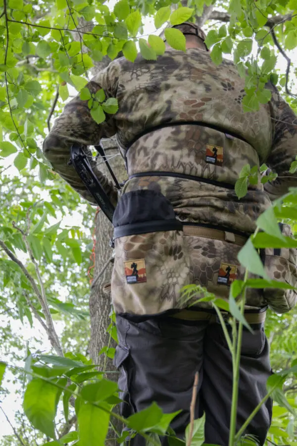 A person dressed in an Ape Canyon Outfitters Lazy Hunter Band Kryptek Highlander is standing on a tree, facing away from the camera. They appear to be engaged in an outdoor activity, surrounded by green foliage. Various equipment is attached to their outfit.