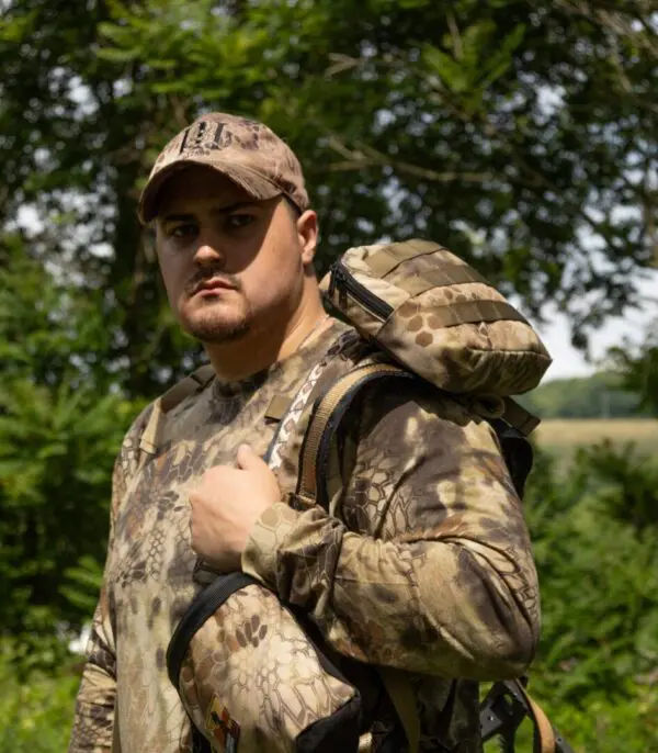 A man wearing camouflage clothing and a cap is outdoors, holding an Ape Canyon Outfitters Climbing Line and Saddle Pouch Combo over his shoulder. He appears to be in a forested area with trees and greenery in the background, facing the camera with a serious expression.