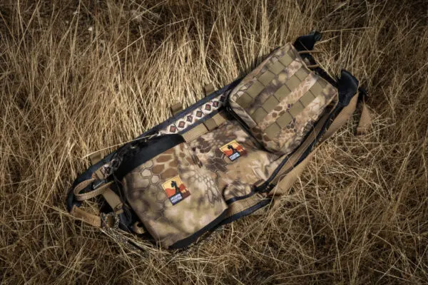 A camouflage-patterned Ape Canyon Outfitters Saddle Pouch Kryptek Highlander lies on dry grass. The backpack features multiple compartments and straps. Two patches with colorful designs are attached to the front. The scene evokes a rugged, outdoor setting.