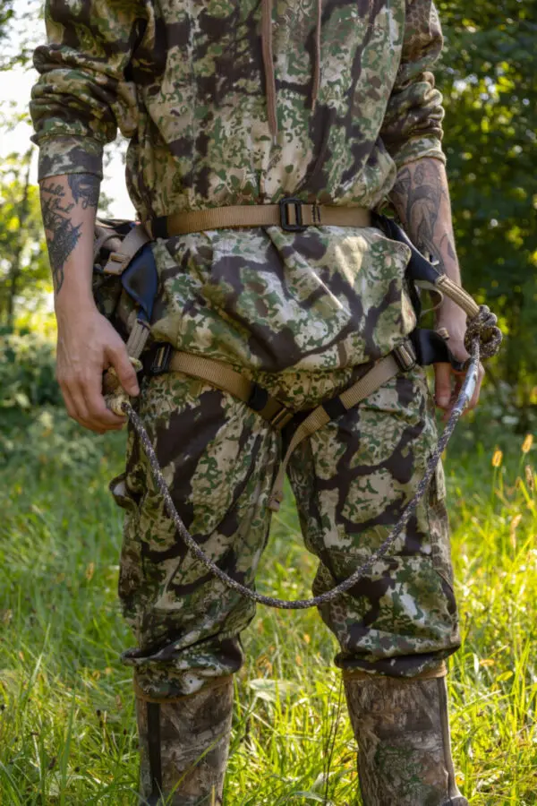 A person stands outdoors wearing camouflage hunting gear, including a jacket, pants, and boots. They are holding a rope and equipped with an Ape Canyon Outfitters Pioneer Saddle Obskura Transitional. The background features greenery and grass, indicating they are in a natural, forested area.