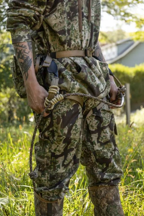 A person wearing camouflage clothing and boots is seen outdoors. They have an Ape Canyon Outfitters Pioneer Saddle Obskura Transitional with ropes and carabiners around their waist and legs, and they are holding onto the harness. Greenery and hints of a building are visible in the background.