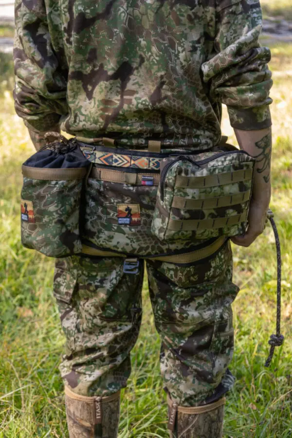 A person wearing camouflage clothing stands outdoors, facing away from the camera. They have various pouches and gear attached to their colorful, patterned Ape Canyon Outfitters Pioneer Saddle Obskura Transitional, suitable for outdoor activities such as hunting or hiking. The surroundings are grassy and natural.