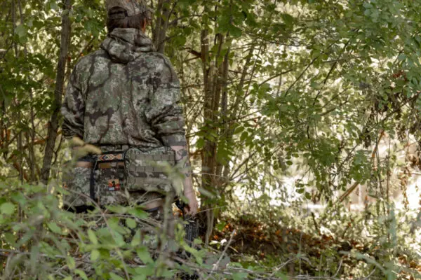A person wearing camouflage clothing and a cap stands with their back turned and holding an Ape Canyon Outfitters One Stick Saddle Pouch Obskura Transitional in the middle of a dense, green forest, partially obscured by foliage.