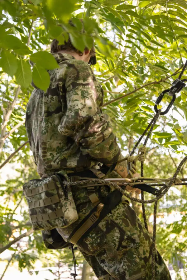 A person dressed in camouflage gear is suspended in a tree, utilizing ropes and harnesses. The foliage around them is dense, making the person blend in with the surroundings. Sunlight filters through the leaves, creating a natural setting while using the Ape Canyon Outfitters Pioneer Saddle Obskura Transitional.