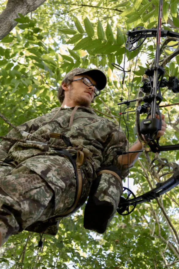 A person dressed in Ape Canyon Outfitters Pioneer Saddle Obskura Transitional clothing is perched in a tree, holding a compound bow. The surroundings are lush with green leaves. The person is looking down, possibly focusing on something below. They are wearing a hat and glasses.
