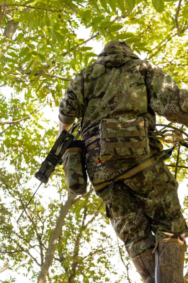 In Ape Canyon Outfitters Pioneer Saddle Obskura Transitional, a person in camouflage clothing, holding a rifle, is looking upward while standing among dense green foliage and trees. The perspective is from below, focusing on their back and the surrounding leaves.