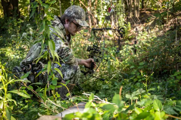 A hunter wearing an Ape Canyon Outfitters Pioneer Saddle Obskura Transitional and a cap is crouched in a forested area, holding a compound bow. Surrounding him are lush green plants and foliage. The scene suggests he is hunting or tracking in the wilderness.
