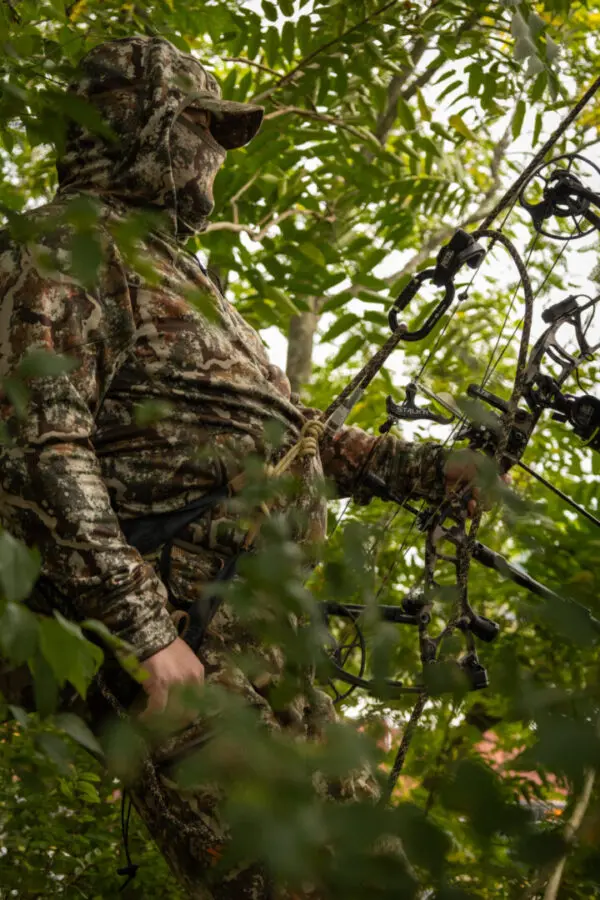 A hunter dressed in an Ape Canyon Outfitters Pioneer Saddle Obskura Transitional stands among dense foliage, holding a compound bow. The surrounding greenery partially obscures the view, blending the hunter into the environment.