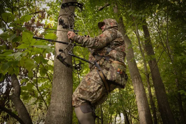 A person dressed in camouflage gear is climbing a tree using an Ape Canyon Outfitters Pioneer Saddle Obskura Transitional. They appear to be preparing for bow hunting, with a compound bow in their hands. The surrounding area is dense with green foliage and trees.