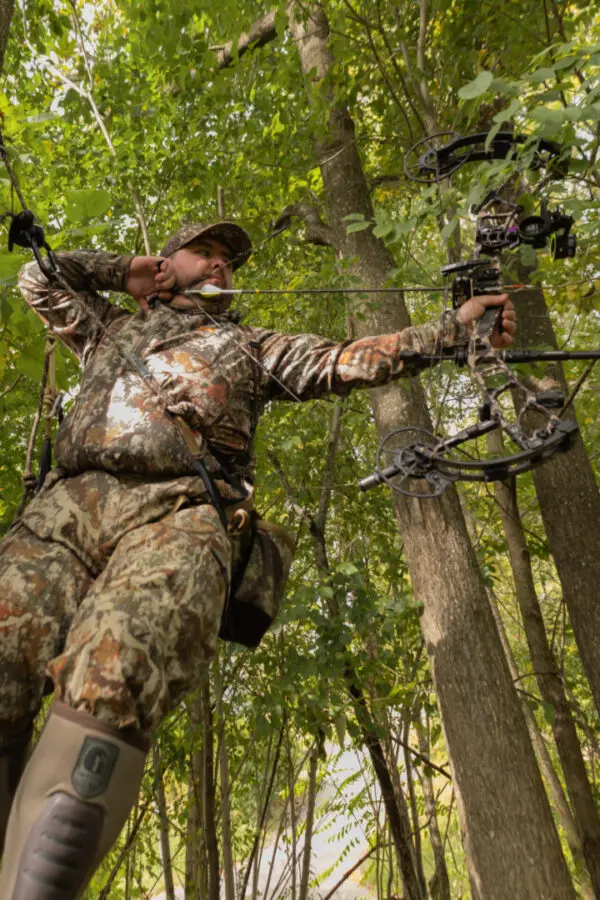 Person dressed in Ape Canyon Outfitters 9.5mm Ghillie, standing among trees, aiming a compound bow. The person appears to be focused, with one hand pulling back the bowstring and the other holding the bow steady while looking at the target.