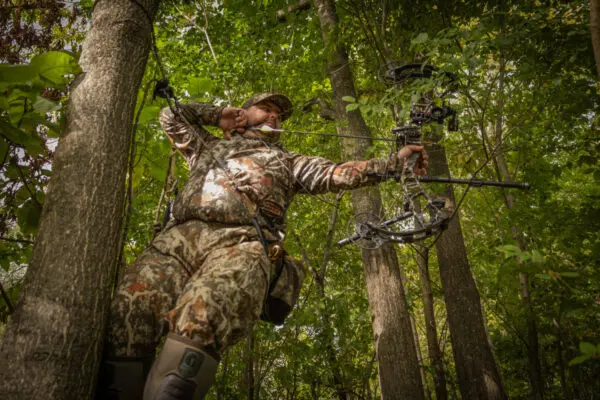 A person dressed in camouflage gear aims a compound bow while standing among trees in a dense forest. The individual is also wearing an Ape Canyon Outfitters Pioneer Saddle Obskura Transitional, boots, and gloves, blending into the woodland environment. Shafts of sunlight filter through the leaves above.