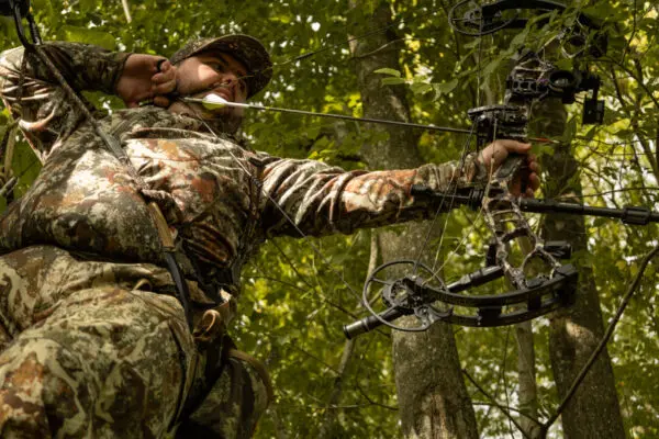 A person in full camouflage gear aims a compound bow upwards, surrounded by dense, leafy trees. The archer appears focused and prepared, likely engaged in a hunting activity while wearing the Ape Canyon Outfitters Pioneer Saddle Obskura Transitional in a forest environment.