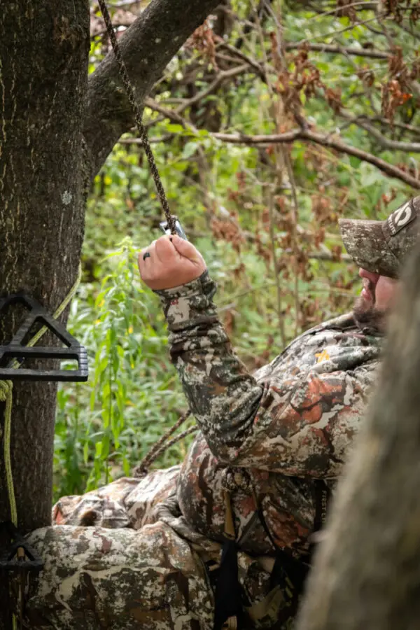 A person in camouflage gear is sitting in a tree and holding onto an Ape Canyon Outfitters 9.5mm Ghillie. They appear to be adjusting or securing something. The surrounding area is dense with green foliage and tree branches.