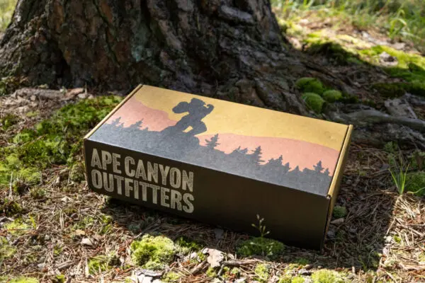 A cardboard box labeled "Ape Canyon Outfitters" rests on mossy ground, leaning against the base of a tree. The box features an illustration of a silhouette of a Bigfoot-like creature against a sunset backdrop with trees.
