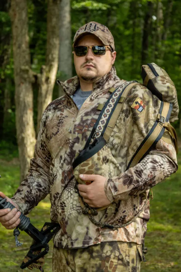 A person stands in a forested area wearing camouflage clothing and sunglasses, holding an Ape Canyon Outfitters Pioneer Saddle Highlander and carrying a backpack.