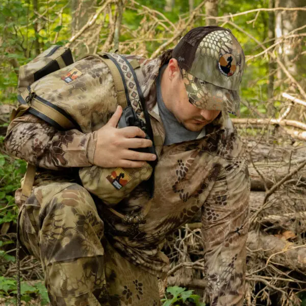 A person in camouflage clothing and an Ape Canyon Outfitters Richardson Trucker Hat crouches in a forest, holding a walkie-talkie. The individual wears a backpack, blending into the woodland surroundings with dense greenery and fallen branches.