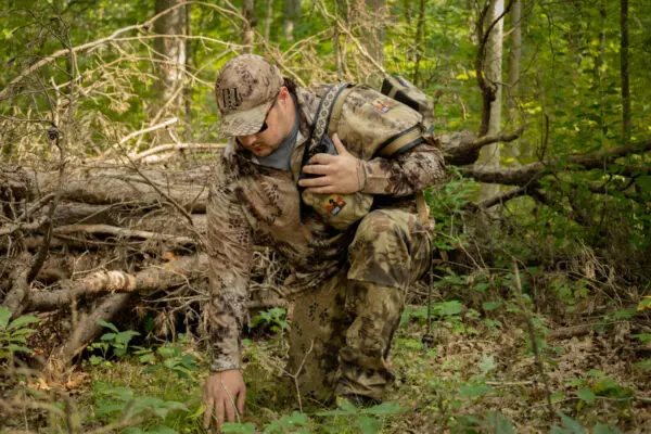 A person in Ape Canyon Outfitters Pioneer Saddle Highlander and a cap kneels in a forest, closely examining the ground. They are surrounded by dense foliage and fallen branches, and appear focused on their task, possibly scouting or tracking.