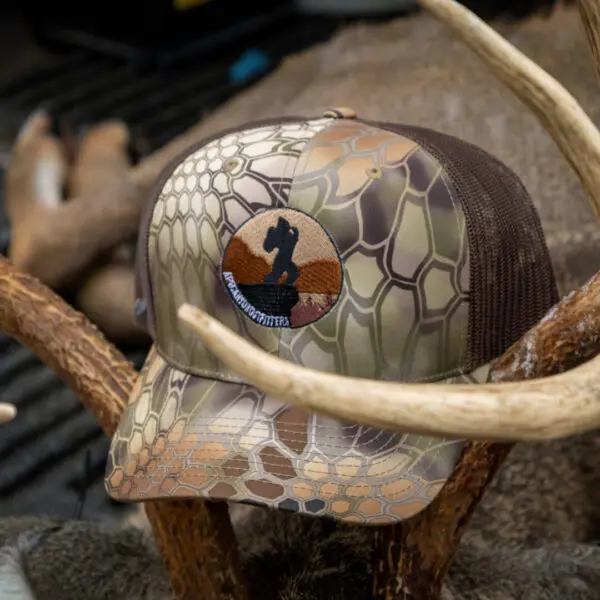An Ape Canyon Outfitters Richardson Trucker Hat with a honeycomb pattern and a round embroidered patch featuring a black silhouette in front of a mountain landscape design is resting on animal antlers. The antlers are displayed in a way that frames the hat.