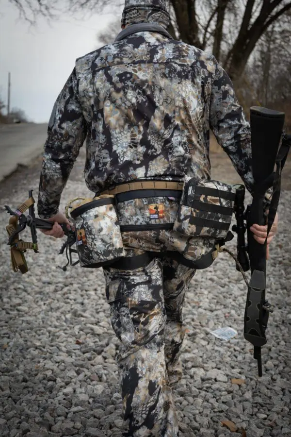 A person in camouflage hunting gear walks on a gravel path, viewed from behind. They carry a compound bow in their left hand and a rifle in their right hand. The person also has a fanny pack and other gear attached to their waist. Trees and a road are in the background.