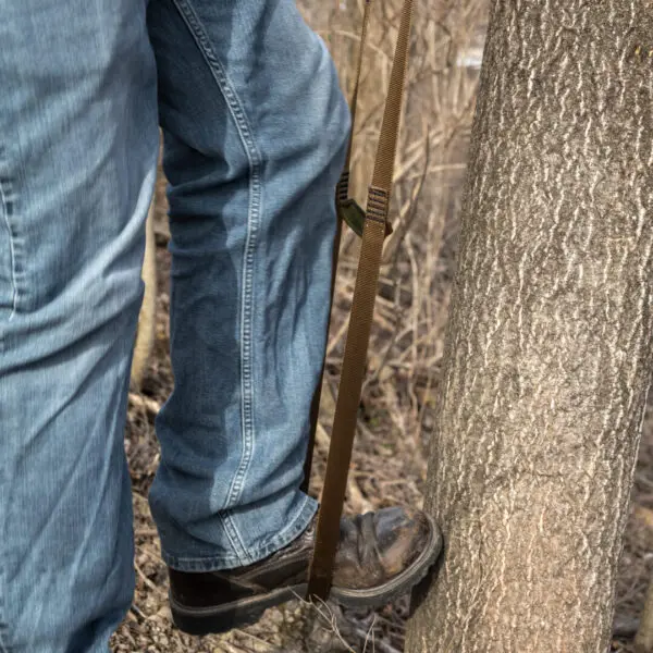 A person wearing blue jeans and black work boots uses an Ape Canyon Outfitters 2 Step Aider attached to a tree trunk. The image focuses on the lower body and the aider tensioned against the tree, indicating climbing or outdoor activity.