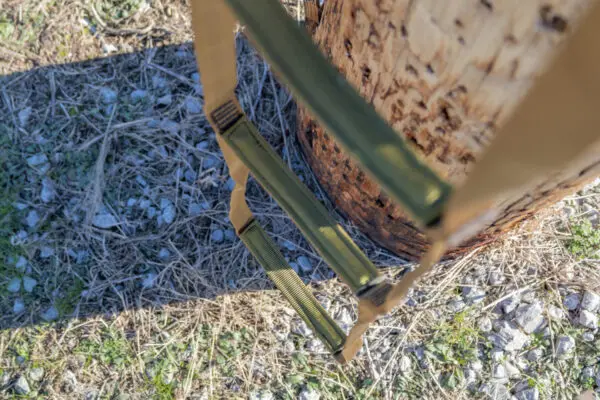 A close-up view of a tree with an Ape Canyon Outfitters 3 Step Aider attached, showing green and beige straps. The ground is covered in dry grass, small rocks, and some scattered greenery. The image was taken in bright daylight, casting shadows.