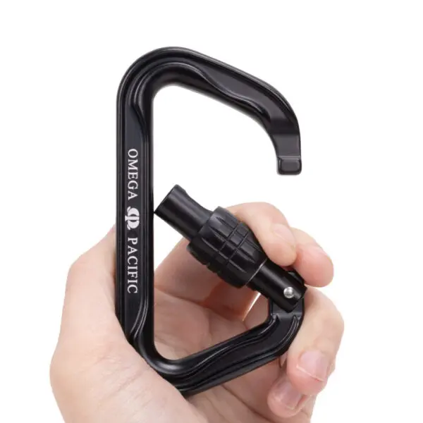A hand holding a black Omega Pacific Standard D Aluminum Keylock Screw-Lok NFPA carabiner with a screw-locking gate mechanism. The fingers grip the carabiner firmly, with the “OMEGA PACIFIC” brand name clearly visible on its side. The background is plain white.
