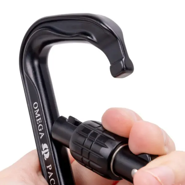 A close-up shot of a hand holding a black Omega Pacific Standard D Aluminum Keylock Screw-Lok NFPA with a twist-lock mechanism. The hand is partially covering the text "Omega" printed on the carabiner, and the twist-lock mechanism is prominently displayed.