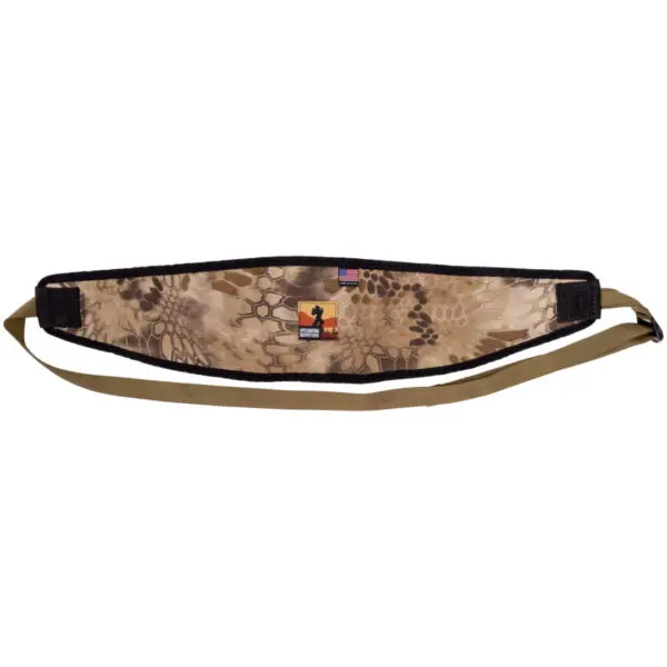 A camouflaged rifle sling featuring a blend of beige and brown patterns. The padded section has a small logo with an American flag design and a silhouette of a person. The Ape Canyon Outfitters Lazy Hunter Band Kryptek Highlander has adjustable straps in a solid, light brown color.