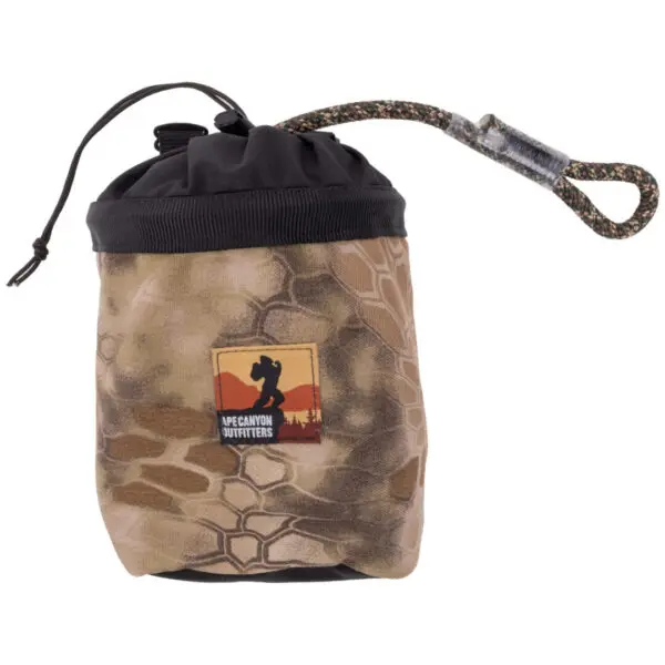 A small, cylindrical, camouflage-patterned pouch with a black drawstring closure and a looped handle. The pouch features a logo of "Ape Canyon Outfitters Saddle Pouch Kryptek Highlander" depicting a silhouette of a hiker against an orange and red mountain background.