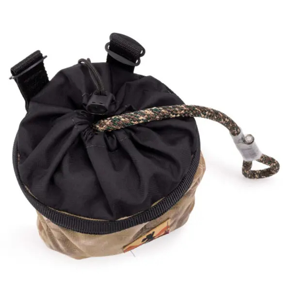 A small, round bag with a drawstring closure is shown. The bag, part of the Ape Canyon Outfitters Climbing Line and Saddle Pouch Combo, is made of camouflage fabric with two black straps and a loop on the top. A braided rope with a secure metal clip is threaded through the drawstring opening.