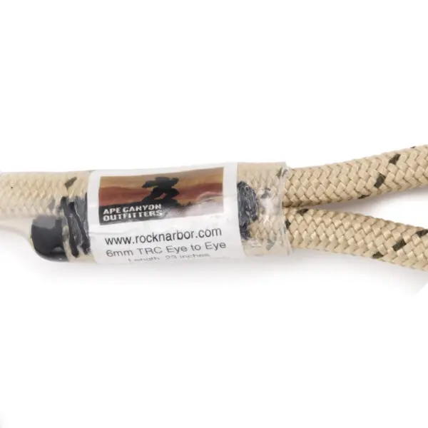 Close-up of a beige braided rope with black speckles, featuring a clear plastic label. The label reads "rocknabor.com Ape Canyon Outfitters 6mm TRC Eye to Eye Hitch Cord 23"" and displays the "Ape Canyon Outfitters" logo. The rope has looped ends secured by black fittings.